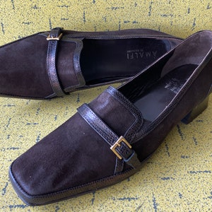 May include: A pair of brown suede heeled loafers with a buckle detail. The loafers are made by the brand "AMALFI di Salerno".