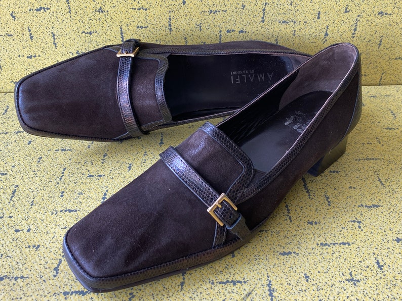 May include: A pair of brown suede heeled loafers with a buckle detail. The loafers are made by the brand "AMALFI di Salerno".