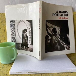 May include: A black and white photo book titled "A Haida Potlatch" by Ulii Stelzer. The book cover features a black and white photo of a person holding a long, carved wooden object. The book is closed and lying on a white surface.
