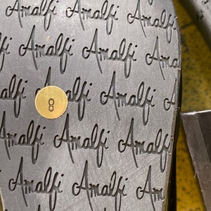 May include: Close-up of the bottom of a black shoe with the word "Amalfi" repeated multiple times. A gold tag with the number "8" is visible.