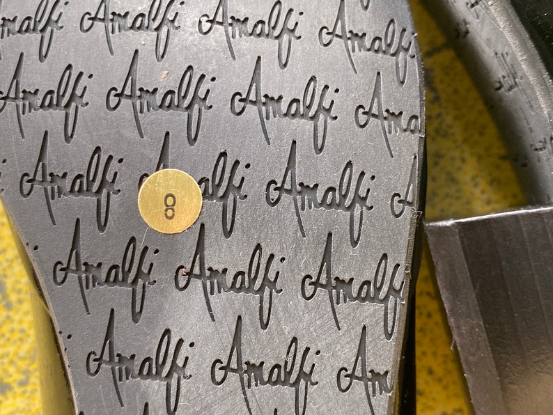 May include: Close-up of the bottom of a black shoe with the word "Amalfi" repeated multiple times. A gold tag with the number "8" is visible.