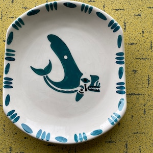 Pode incluir: Um prato de cerâmica branco com uma borda azul-esverdeada e um design de baleia azul-esverdeada estilizado no centro.