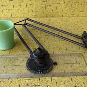 May include: A black drafting tool with a protractor and adjustable arms. The tool is on a clear plastic ruler with markings in inches.