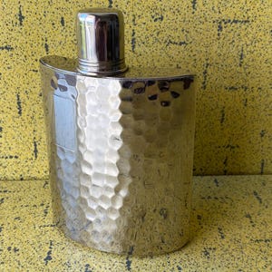 May include: A silver-coloured flask with a hammered texture and a shiny, cylindrical cap. The flask has a curved, rectangular shape and is designed to hold liquids. The background is a textured yellow surface.