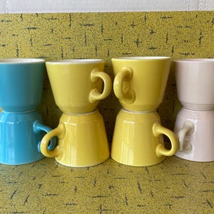 Może przedstawiać: Cztery ceramiczne kubki w odcieniach niebieskiego, żółtego i różowego ułożone na żółtej powierzchni. Każdy kubek ma zaokrąglony uchwyt i kontrastujące białe wnętrze. Kubki są ułożone parami, tworząc atrakcyjną wizualnie ekspozycję.