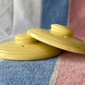 Op de afbeelding: Twee gele keramische deksels met een verhoogde knop aan de bovenkant. De deksels hebben een geribbeld ontwerp en een klein gaatje in het midden.