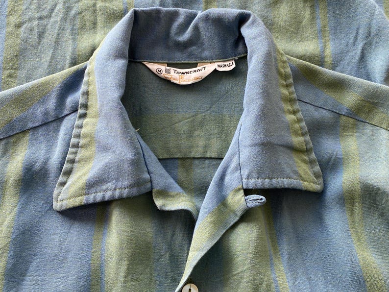 May include: Close-up of a blue and green striped button-down shirt. The shirt has a classic collar and a button closure. The tag reads "TOWN CRAFT WASHABLE". The fabric has a soft, textured appearance.