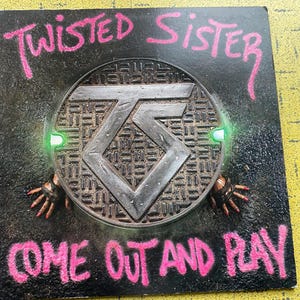 Può includere: Copertina dell'album "Come Out and Play" dei Twisted Sister. L'artwork presenta un tombino metallico con il logo della band, circondato da testo in stile graffiti in rosa. Lo sfondo è nero con un aspetto strutturato.