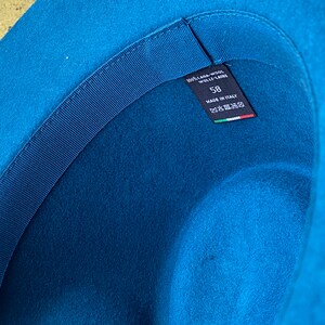 May include: The inside of a blue felt hat with a black ribbon and a label that reads "100% Lama Wool - Wolle - Laine" and "58" with "Made in Italy" and an Italian flag.