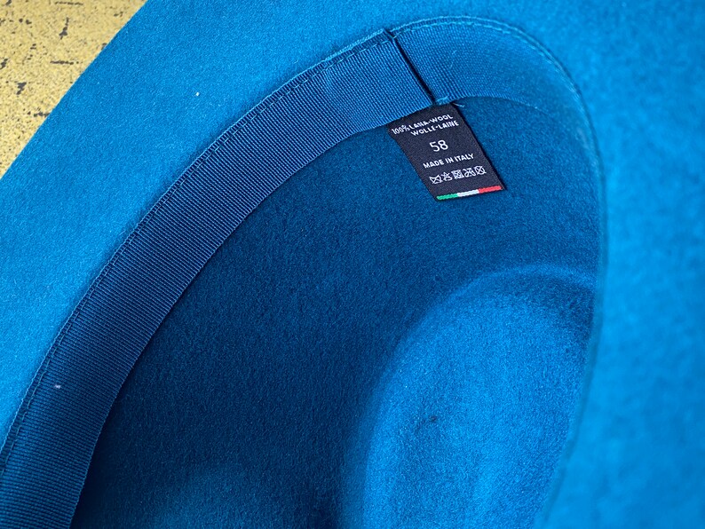 May include: The inside of a blue felt hat with a black ribbon and a label that reads "100% Lama Wool - Wolle - Laine" and "58" with "Made in Italy" and an Italian flag.