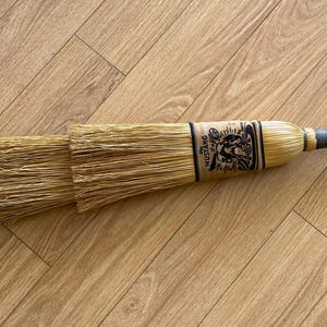 May include: A straw broom with a tan handle and a tan band featuring a black graphic of a horse and the text "The MUSTANG". The broom head consists of layered, light-coloured natural fibres bound together at the top with a dark-coloured wrap.