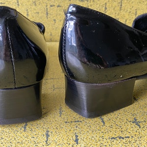 May include: A pair of black patent leather heeled shoes with a low block heel. The shoes have a simple design and are made of a smooth, shiny material.