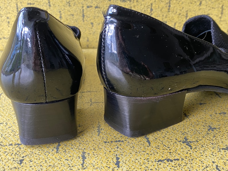May include: A pair of black patent leather heeled shoes with a low block heel. The shoes have a simple design and are made of a smooth, shiny material.