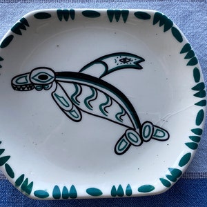 Pode incluir: Um prato de cerâmica branco com uma borda verde e um desenho estilizado verde e preto de uma criatura marinha com um olho nas costas.