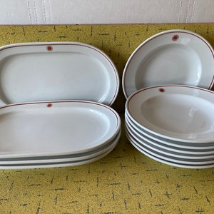 Può includere: Un set di piatti ovali e rotondi in ceramica bianca con un logo rosso. I piatti sono impilati e disposti su una superficie con un motivo giallo.