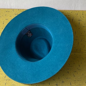 May include: A teal blue felt fedora hat with a wide brim. The hat has a black ribbon around the crown.
