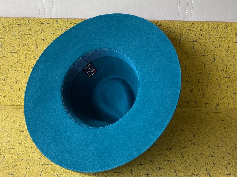 May include: A teal blue felt fedora hat with a wide brim. The hat has a black ribbon around the crown.