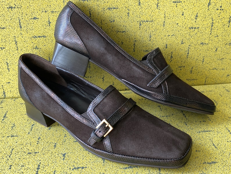 May include: A pair of brown suede heeled loafers with a buckle detail. The loafers have a pointed toe and a block heel.
