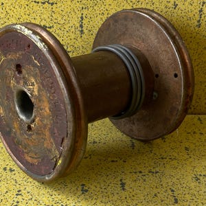Op de afbeelding: Een grote houten spoel met metalen uiteinden. De spoel is roestig en heeft een grijs elastiek eromheen gewikkeld. De spoel ligt op een geel oppervlak.