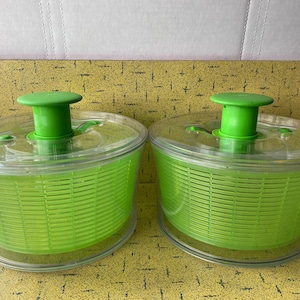 Op de afbeelding: Twee transparante plastic slacentrifuges met felgroene manden en deksels. Elke centrifuge heeft een groene handgreep aan de bovenkant. De centrifuges staan op een gele ondergrond.