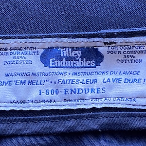 May include: A close-up shot of a white label with blue text and a blue border, stitched onto dark blue fabric. The label reads "Tilley Endurables" and includes washing instructions and fabric composition details.