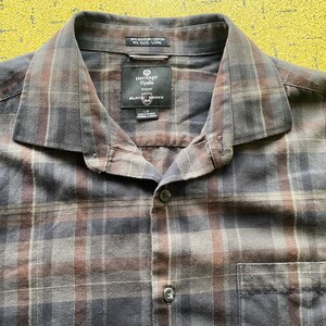 May include: A checked shirt in brown, grey, and blue tones. The shirt features a collar, a chest pocket, and a label reading "Heritage Vivella Black Brown". It is made from a blend of cotton and wool. The shirt has a button-down design.