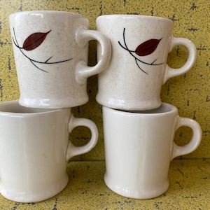 Può includere: Set di quattro tazze in ceramica bianca con motivi di foglie marroni. Le tazze hanno una finitura macchiettata e sono impilate l'una sull'altra.