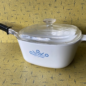 以下が含まれることがあります： 青い花柄の白い正方形のキャセロール皿と透明なガラスの蓋。皿には黒いハンドルが付いています。