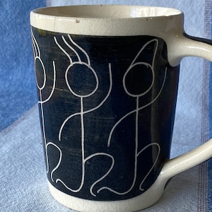 Pode incluir: Uma caneca de cerâmica branca com um desenho preto e branco de figuras de palitos. As figuras estão dançando e têm um toque caprichoso.