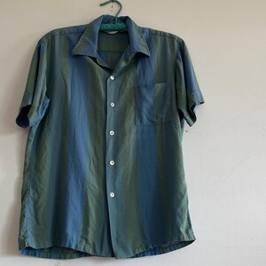 May include: A short-sleeved button-down shirt in shades of teal and blue, hanging on a green hanger. The shirt has a chest pocket and a classic collar. The fabric appears to have a subtle sheen.