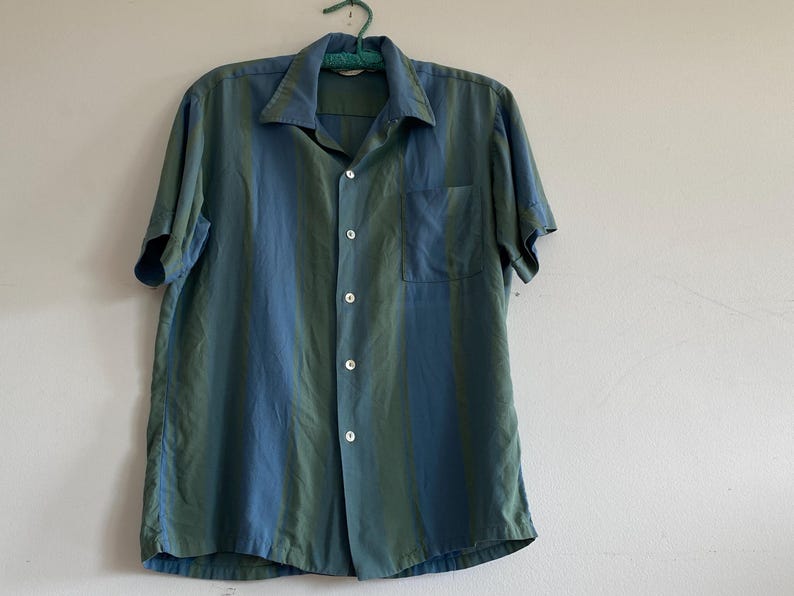 May include: A short-sleeved button-down shirt in shades of teal and blue, hanging on a green hanger. The shirt has a chest pocket and a classic collar. The fabric appears to have a subtle sheen.