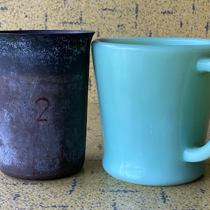 May include: A vintage metal measuring cup with the number "2" marked on the side, next to a light green ceramic mug with a handle.