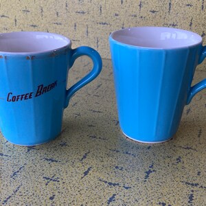 Peut inclure: Deux tasses à café en céramique bleu clair avec un design côtelé. L'une des tasses porte l'inscription "Coffee Break" en lettres rouges. Les deux tasses ont un intérieur blanc et une anse incurvée.