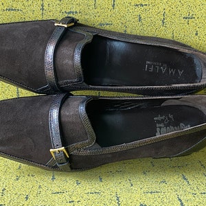 May include: A pair of brown suede loafers with a buckle closure. The loafers are by the brand Amalfi by Randolph.