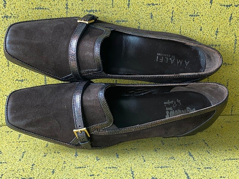May include: A pair of brown suede loafers with a buckle closure. The loafers are by the brand Amalfi by Randolph.