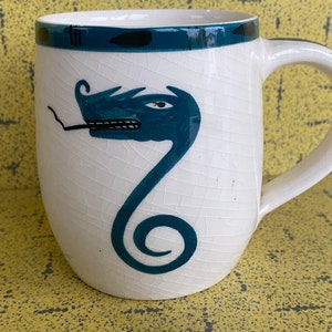 Pode incluir: Caneca de cerâmica branca com uma borda azul e preta. A caneca apresenta um desenho pintado à mão de um dragão verde com uma cauda enrolada.