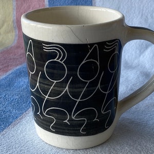 Pode incluir: Caneca de cerâmica branca com um desenho em preto e branco de figuras estilizadas. As figuras são delineadas em branco e têm corpos e cabeças pretos.