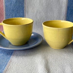 Op de afbeelding: Twee gele theekopjes met een blauwe schotel. De kopjes hebben een geribbeld ontwerp en zijn gemaakt van keramiek.