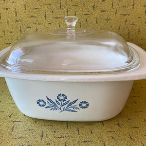 Może przedstawiać: Biała ceramiczna naczynie do zapiekania z szklaną pokrywką i niebieskim wzorem kwiatowym. Naczynie ma zaokrąglony kształt i szeroką krawędź.