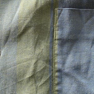 May include: Close-up of a blue and green striped fabric, possibly linen or cotton. The fabric has a textured appearance with a button and a pocket detail visible. The button is off-white. The pocket is on the right side.