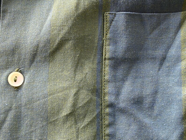 May include: Close-up of a blue and green striped fabric, possibly linen or cotton. The fabric has a textured appearance with a button and a pocket detail visible. The button is off-white. The pocket is on the right side.