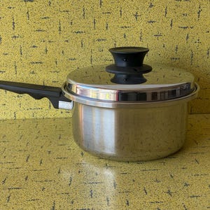 Può includere: Casseruola in acciaio inossidabile con coperchio e manico nero. Il coperchio ha un pomello nero. La pentola è appoggiata su una superficie gialla.