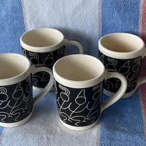 Pode incluir: Quatro canecas de cerâmica branca com alças pretas e uma faixa preta ao redor da parte superior. A faixa preta apresenta um desenho branco de figuras estilizadas.