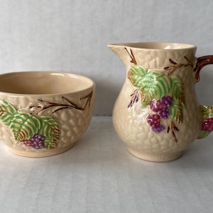 May include: A cream-colored ceramic sugar bowl and creamer set with a berry and leaf design. The set has a textured surface and is decorated with green leaves, brown branches, and purple berries.