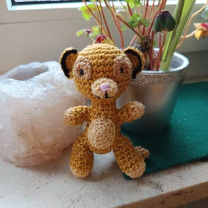 Könnte beinhalten: Ein gehäkeltes Stofftier-Löwenjunges mit einem hellbraunen Körper und einem dunkelbraunen Gesicht. Das Löwenjunges hat schwarze Ohren und eine rosa Nase und einen rosa Mund.