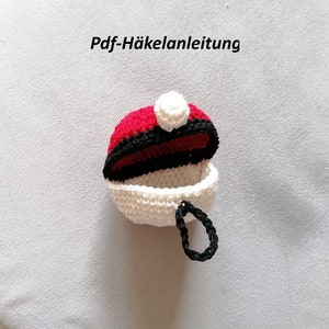 Op de afbeelding: Een gehaakte rode, zwarte en witte tas met een witte pompon bovenop. De tas is open en heeft een zwart gehaakt lusje als handvat. De tekst "Pdf-Häkelanleitung" is zichtbaar boven de tas.