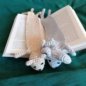Puede incluir: Dos marcapáginas de animales de crochet, uno beige y otro gris, con ojos negros. Los marcapáginas tienen forma de animales con orejas largas y están sobre un libro.