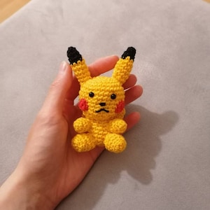 Op de afbeelding: Een gehaakt geel Pikachu knuffel met zwarte oren en rode wangen. Het speelgoed wordt in de hand van een persoon gehouden.