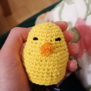 Könnte beinhalten: Ein handgehäkeltes, gelbes Küken mit schwarzen Augen und einem orangefarbenen Schnabel. Das Küken hat eine eiförmige Gestalt und scheint ein kleines Spielzeug oder Dekorationsartikel zu sein. Die Häkelstiche sind sichtbar und verleihen ihm eine strukturierte Optik.