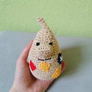 Puede incluir: Una muñeca de crochet beige con una nariz larga y una cara con parches coloridos. La muñeca está siendo sostenida en una mano.
